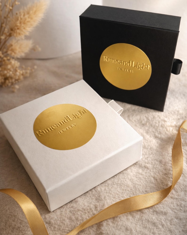May include: Two jewelry boxes, one black and one white, with gold foil labels. The labels read "RuneandLight JEWELRY". A gold ribbon is draped across the surface. The boxes are closed, suggesting they contain jewelry.