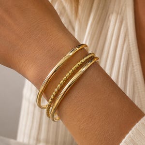 Może przedstawiać: Zestaw trzech złotych bransoletek typu bangle, składający się z dwóch gładkich, polerowanych okrągłych obręczy oraz jednej środkowej obręczy o fakturze skręconego sznura. Bransoletki noszone są razem, prezentując odblaskowe wykończenie.