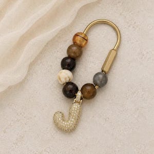 May include: A gold-toned metal keychain featuring a U-shaped bar and a series of round beads in shades of brown, cream, and grey. A gold-toned letter J charm encrusted with small clear stones hangs from the bottom of the beaded strand.