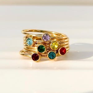May include: A stack of thin gold-toned metal rings, each featuring a single small round gemstone in various colours including red, green, yellow, light blue, and lavender. The rings are arranged in a cluster, showcasing the polished gold bands.
