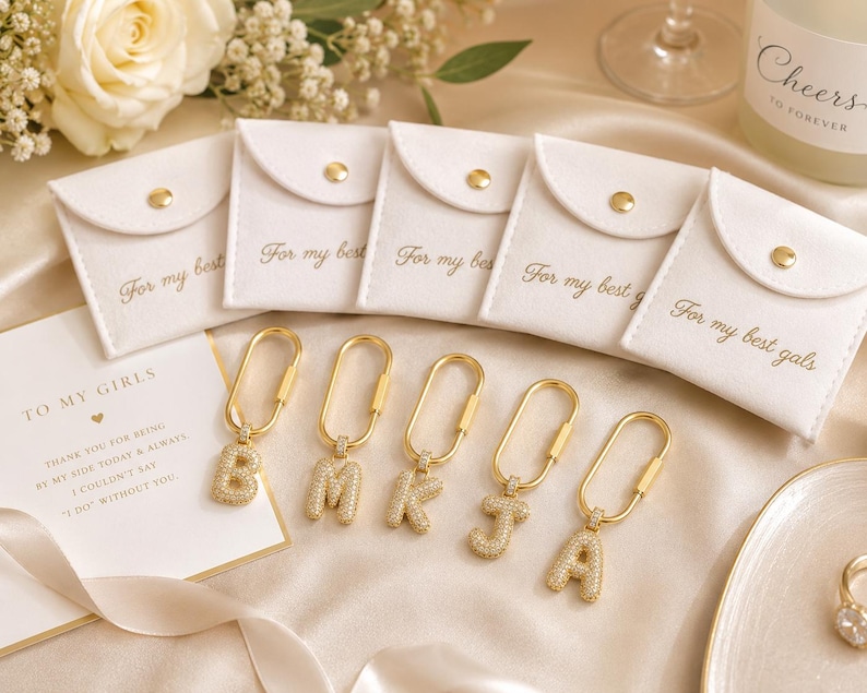 Op de afbeelding: Gouden initialen sleutelhangers met sprankelende kristallen accenten, gepresenteerd met witte zakjes en een kaart met de tekst "To My Girls". De zakjes zijn voorzien van de tekst "For my best" en "For my best gals".