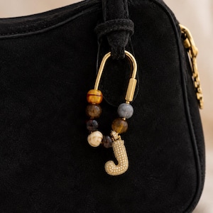 May include: A black suede handbag with a gold-colored clasp and chain detail. A gold-colored charm with the letter "J" encrusted with small, sparkling stones hangs from the handle. The charm is attached to a gold-colored carabiner with a beaded accent.