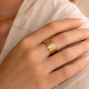 May include: A gold-colored ring with a floral pattern. The ring is wide and wraps around the finger. The floral design is etched into the metal, creating a textured surface. The ring is worn on a hand.