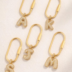 Op de afbeelding: Goudkleurige sleutelhangers met de letters A, K, J, S en M. Elke letter is bezet met kleine, sprankelende kristallen. De sleutelhangers hebben een eenvoudig, ovaalvormig ontwerp.