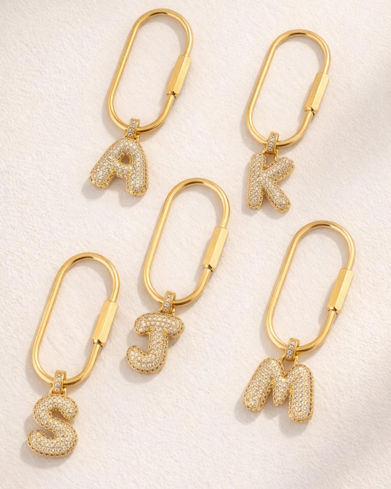 Op de afbeelding: Goudkleurige sleutelhangers met de letters A, K, J, S en M. Elke letter is bezet met kleine, sprankelende kristallen. De sleutelhangers hebben een eenvoudig, ovaalvormig ontwerp.