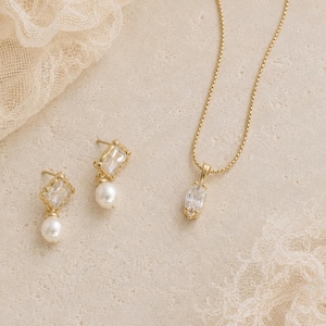 May include: A gold-toned jewelry set featuring a necklace and earrings. The necklace has a delicate chain with a pendant. The earrings are stud style with a square-cut clear stone and a dangling pearl. The jewelry is displayed on a neutral surface.