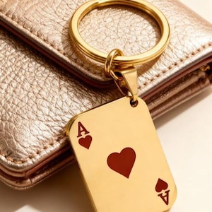 May include: Gold-colored keychain with a rectangular pendant featuring the Ace of Hearts playing card design. The keychain is attached to a gold ring, and the background shows a gold-colored wallet.