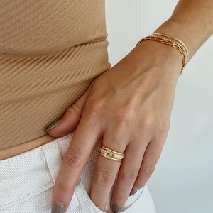 Puede incluir: Un anillo de oro con una banda texturizada y un peque&ntilde;o dise&ntilde;o en relieve en la parte superior. El anillo se lleva en el dedo &iacute;ndice de una mano.