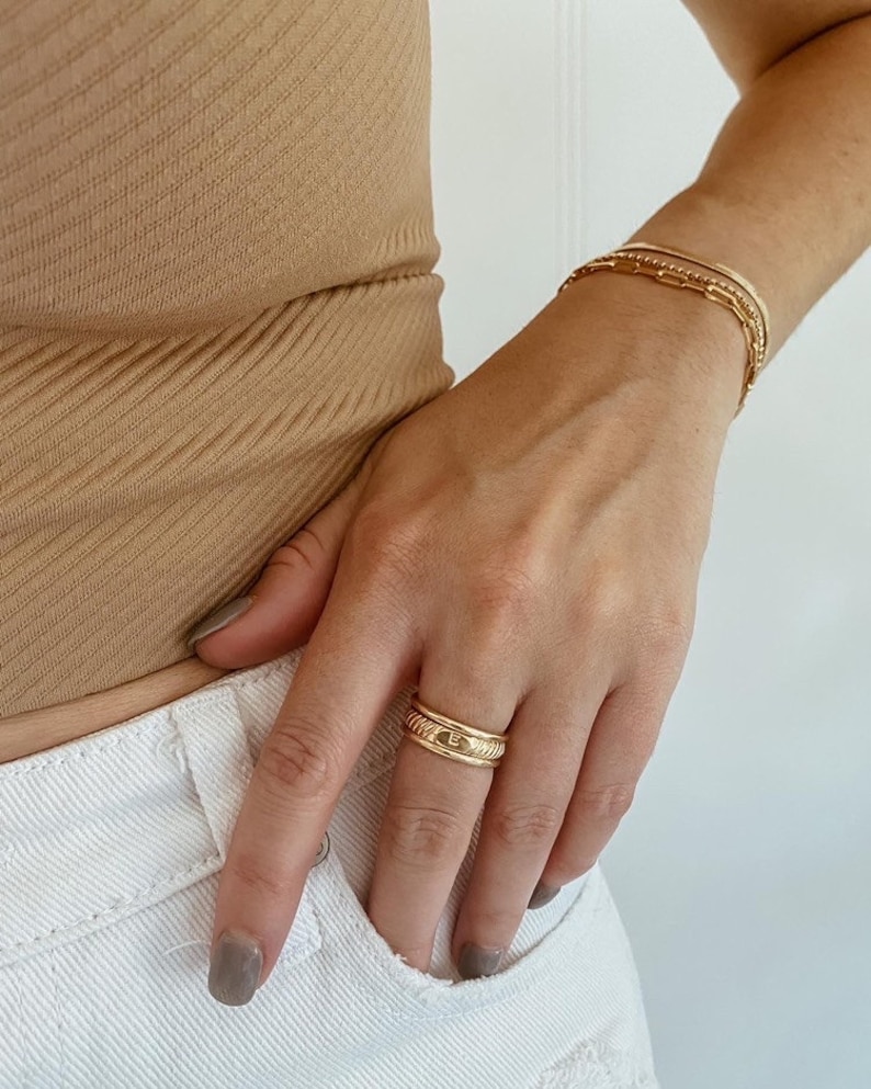 Puede incluir: Un anillo de oro con una banda texturizada y un peque&ntilde;o dise&ntilde;o en relieve en la parte superior. El anillo se lleva en el dedo &iacute;ndice de una mano.