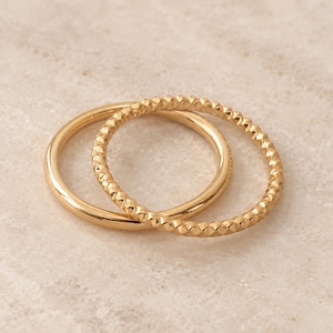 May include: Two gold rings are interlocked. One ring features a smooth, polished surface, while the other ring has a textured, faceted surface with a repeating geometric pattern. Both rings are thin, circular bands made of a shiny, metallic material.