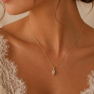 May include: A delicate gold necklace with a marquise-cut pendant. The pendant is a clear, sparkling gemstone. The necklace is worn on a person's neck, with a lace garment visible on the shoulders.