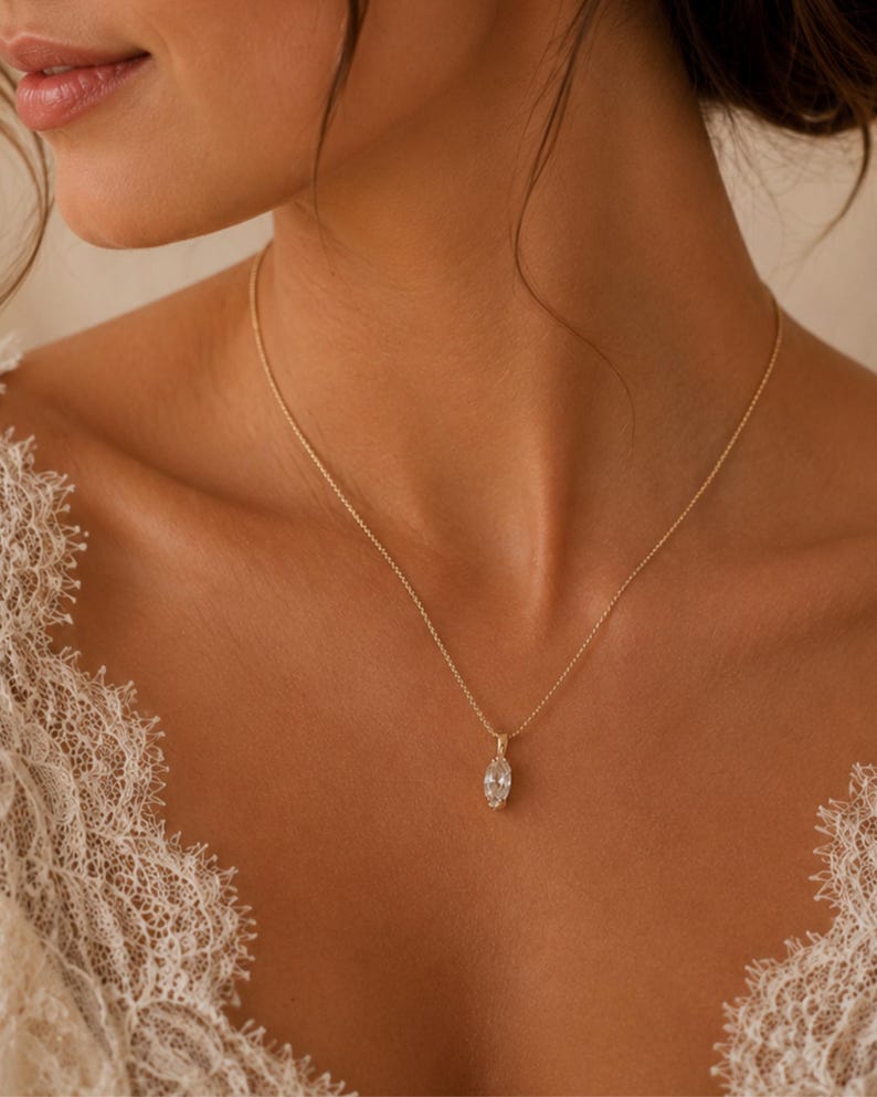 May include: A delicate gold necklace with a marquise-cut pendant. The pendant is a clear, sparkling gemstone. The necklace is worn on a person's neck, with a lace garment visible on the shoulders.