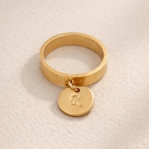 May include: A gold-colored ring with a flat band and a circular charm. The charm is engraved with the letter "R". The ring and charm are made of a shiny, metallic material.