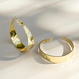 May include: Two gold-colored hoop earrings with a hammered texture. The earrings are on a white surface, with shadows cast across the surface. The earrings are a simple, elegant design.