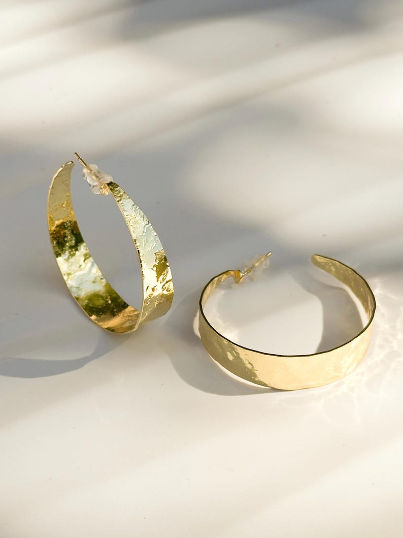 May include: Two gold-colored hoop earrings with a hammered texture. The earrings are on a white surface, with shadows cast across the surface. The earrings are a simple, elegant design.