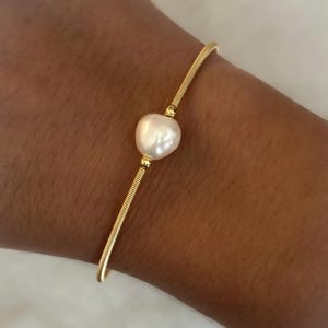 May include: A gold-colored bracelet featuring a large, off-white pearl as its focal point. The bracelet has a delicate, minimalist design with a textured band and smooth, curved ends. The pearl is centered on the bracelet.