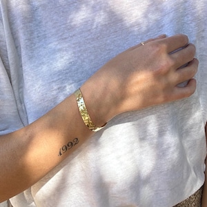 May include: A gold-coloured cuff bracelet with a textured surface. The bracelet is worn on the wrist of a person wearing a white t-shirt. The year "1992" is tattooed on the person's arm.