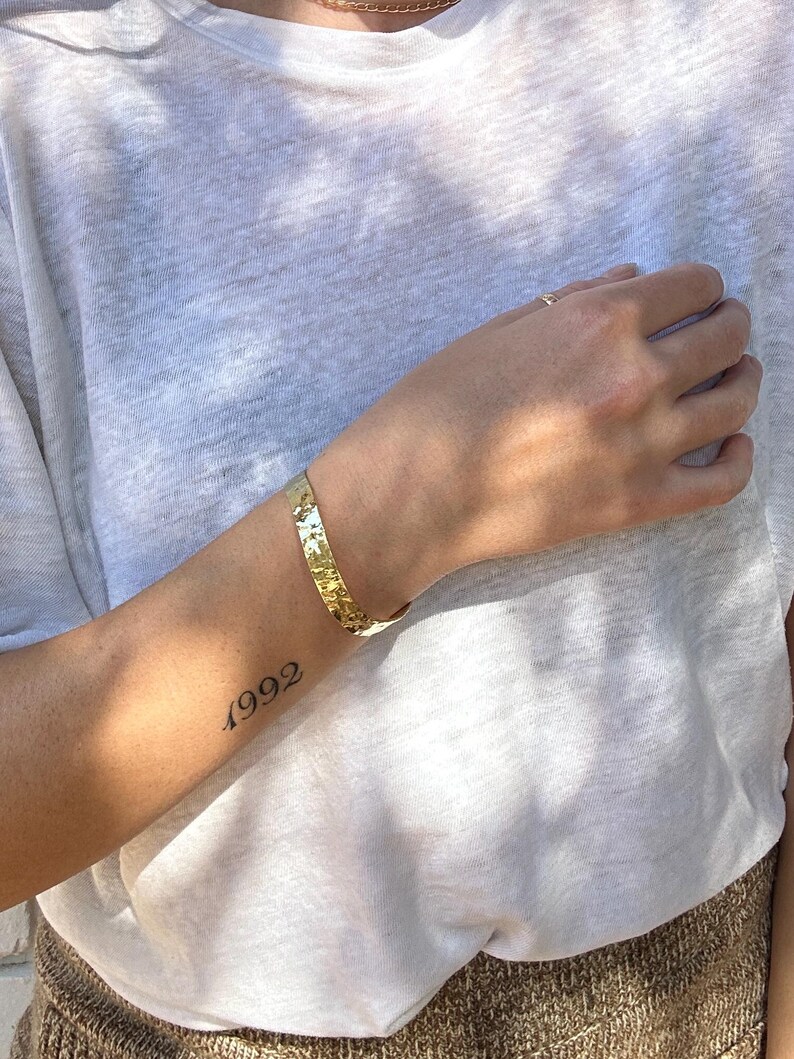 May include: A gold-coloured cuff bracelet with a textured surface. The bracelet is worn on the wrist of a person wearing a white t-shirt. The year "1992" is tattooed on the person's arm.