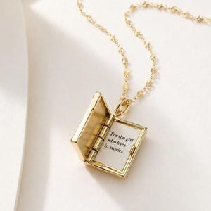 May include: A gold-toned metal pendant necklace shaped like an open book. The interior page features the black printed text: For the girl who lives in stories. The pendant hangs from a delicate gold-toned chain with small, textured, circular links.