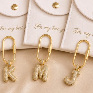 Op de afbeelding: Goudkleurige sleutelhangers met de letters K, M en J, elk ingelegd met kleine, heldere steentjes. De sleutelhangers hangen aan een gouden sluiting. De achtergrond toont witte zakjes met de tekst "For my best".