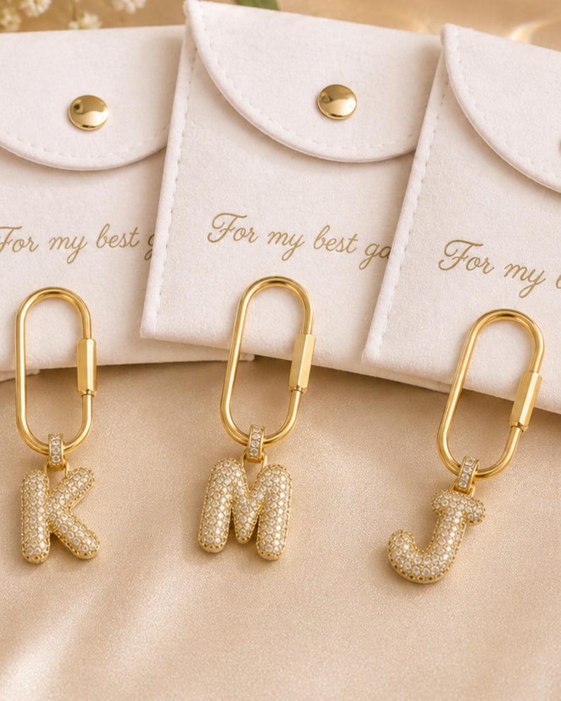 Op de afbeelding: Goudkleurige sleutelhangers met de letters K, M en J, elk ingelegd met kleine, heldere steentjes. De sleutelhangers hangen aan een gouden sluiting. De achtergrond toont witte zakjes met de tekst "For my best".