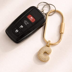 Op de afbeelding: Een zwarte autosleutelhanger en een goudkleurige sleutelhanger met een 'C'-hanger. De hanger is bezet met kleine, glinsterende kristallen. De sleutelhanger heeft een karabijnhaaksluiting.