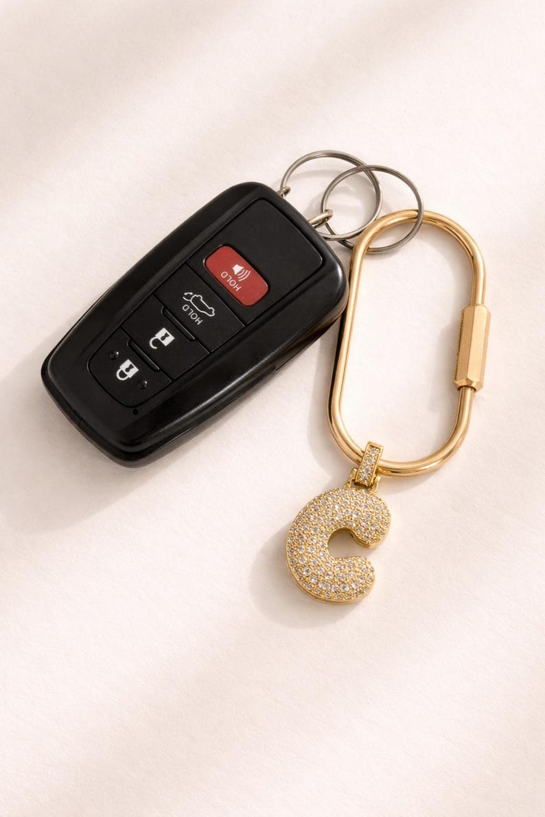 Op de afbeelding: Een zwarte autosleutelhanger en een goudkleurige sleutelhanger met een 'C'-hanger. De hanger is bezet met kleine, glinsterende kristallen. De sleutelhanger heeft een karabijnhaaksluiting.