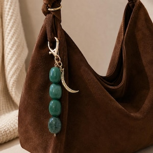 May include: A dark brown suede hobo bag featuring a gold-toned metal lobster clasp charm. The charm consists of four smooth, round, dark green beads stacked vertically and a small, textured gold-toned crescent moon pendant attached to the side.