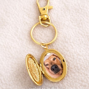 May include: Gold-colored locket keychain with a photo of a dog inside. The locket is oval-shaped and opens to reveal the photo. The keychain has a clasp and a ring. Text on the image reads: "option to include small photo inside locket."