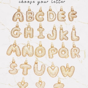Op de afbeelding: Gouden letterhangers, elk bezet met kleine, glinsterende steentjes. De hangers worden getoond tegen een achtergrond met marmer-effect. De tekst "choose your letter" staat er in sierlijke letters boven.