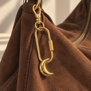 May include: A brown suede handbag featuring a gold-toned metal lobster clasp attached to a rectangular carabiner link. A polished gold crescent moon charm hangs from the bottom of the carabiner, which is secured to the bag by a matching gold ring.