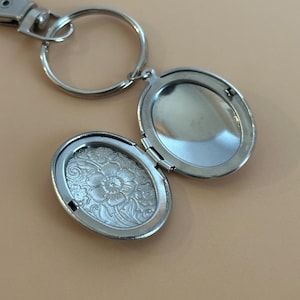 May include: Silver-colored oval locket keychain, open to reveal a floral embossed design on one side. The locket is attached to a key ring and a clasp. The background is a solid tan color.