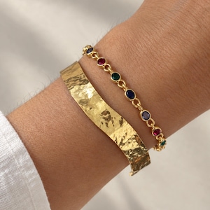Puede incluir: Un brazalete rígido dorado con superficie martillada y texturizada, junto a una pulsera de cadena dorada con pequeñas piedras preciosas redondas y multicolores en tonos rojo, verde y azul, con un diseño de eslabones metálicos.
