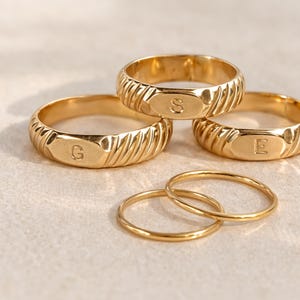 May include: A collection of gold-colored rings. Several rings have a ribbed design and feature engraved letters: G, S, and E. There are also thinner, plain gold bands. The rings are displayed on a light-colored surface.