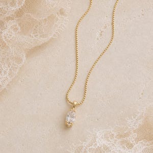 May include: A gold-colored necklace with a delicate chain and a pendant featuring a clear, oval-cut gemstone. The necklace is displayed on a textured, off-white surface with lace accents, creating an elegant and refined aesthetic.
