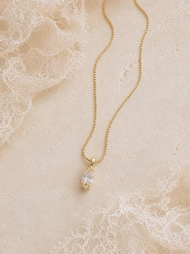 May include: A gold-colored necklace with a delicate chain and a pendant featuring a clear, oval-cut gemstone. The necklace is displayed on a textured, off-white surface with lace accents, creating an elegant and refined aesthetic.