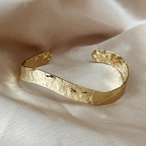 May include: A gold-coloured cuff bracelet with a hammered texture. The open-ended bracelet has a slightly curved shape and rests on a light-coloured, textured surface. The bracelet is a piece of jewellery.