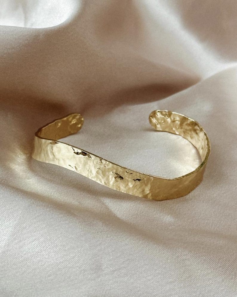 May include: A gold-coloured cuff bracelet with a hammered texture. The open-ended bracelet has a slightly curved shape and rests on a light-coloured, textured surface. The bracelet is a piece of jewellery.