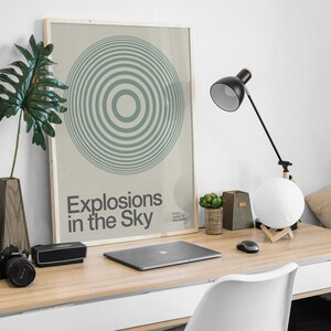 May include: A framed poster with a green and white concentric circle design. The poster has the text "Explosions in the Sky" and the band's name and location. The poster is hanging on a white wall above a desk with a laptop, a lamp, and a moon lamp.
