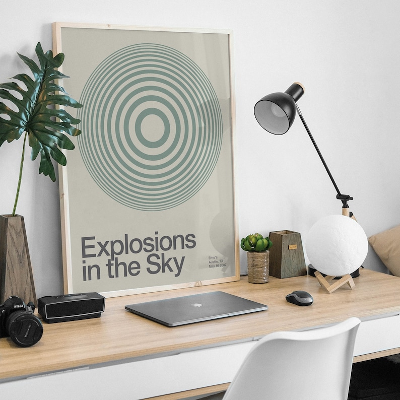 May include: A framed poster with a green and white concentric circle design. The poster has the text "Explosions in the Sky" and the band's name and location. The poster is hanging on a white wall above a desk with a laptop, a lamp, and a moon lamp.