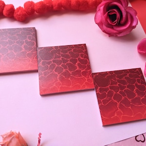 May include: Three red square sticky notes with a gradient pattern of red and dark red. The notes have a textured surface with a pattern of small circles. The notes are arranged in a row on a pink surface.