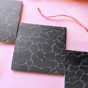 May include: Three black square sticky notes with a white abstract pattern of circles and lines. The notes are on a pink surface.
