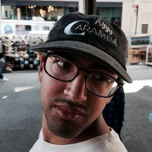 Puede incluir: Una persona con una gorra negra con el texto "Aramba" en blanco y azul claro. La persona también lleva gafas con montura oscura y una camiseta blanca.