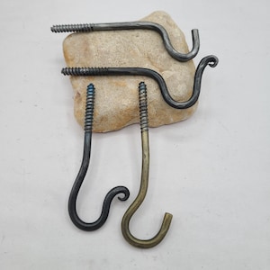 May include: Four handcrafted metal screw-in hooks in various finishes: dark grey, black, and brass. The hooks are displayed on a light brown stone, showcasing their unique, curved designs. The screw threads are visible, highlighting the rustic, industrial aesthetic.