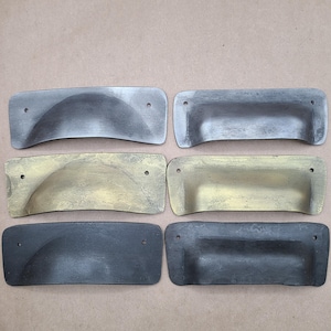 May include: Six cabinet pulls in various finishes. The pulls are made of metal and have a curved shape. The finishes include black, silver, and gold.