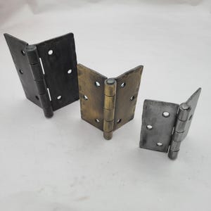 May include: Three metal door hinges in varying sizes and finishes. The hinges are in black, bronze, and silver tones. Each hinge has a V-shape with multiple screw holes and a central hinge pin. The hinges are likely for furniture or doors.
