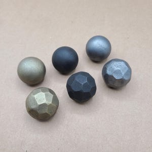 May include: Six metal spheres in various finishes: bronze, black, grey, and faceted dark grey. The spheres are arranged on a neutral tan surface, showcasing their different textures and colours. These decorative spheres could be used as cabinet knobs or decorative accents.