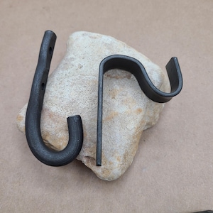 May include: Two black metal hooks, one with a curved shape and the other with a straight, angled shape. The hooks are resting on a light brown rock.