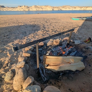 May include: A campfire with a log burning in the middle of a circle of rocks on a sandy beach. The fire is surrounded by a metal grate. The beach is next to a body of water with mountains in the background.