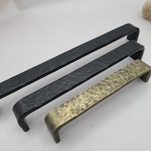 May include: Three metal cabinet pulls in different lengths and finishes. The top pull is black, the middle pull is dark grey, and the bottom pull is brass. All have a hammered texture and a curved, U-shaped design.